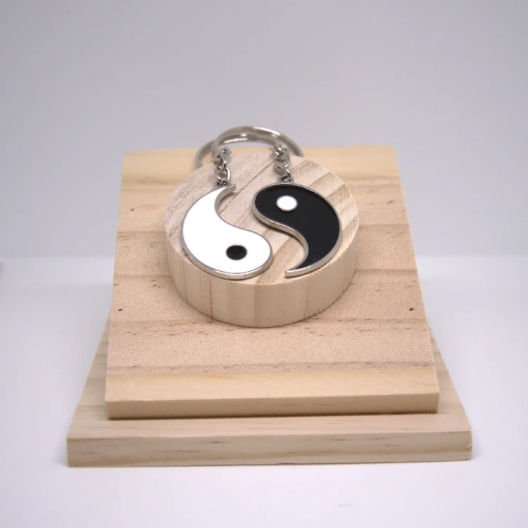 Zestaw breloczków Yin Yang ze stali nierdzewnej na drewnianej podstawce, dwa elementy tworzące symbol harmonii i połączenia.